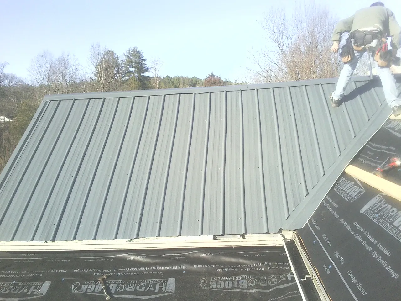 Crew installing dark green standing seam panels over synthetic underlayment in Boswell