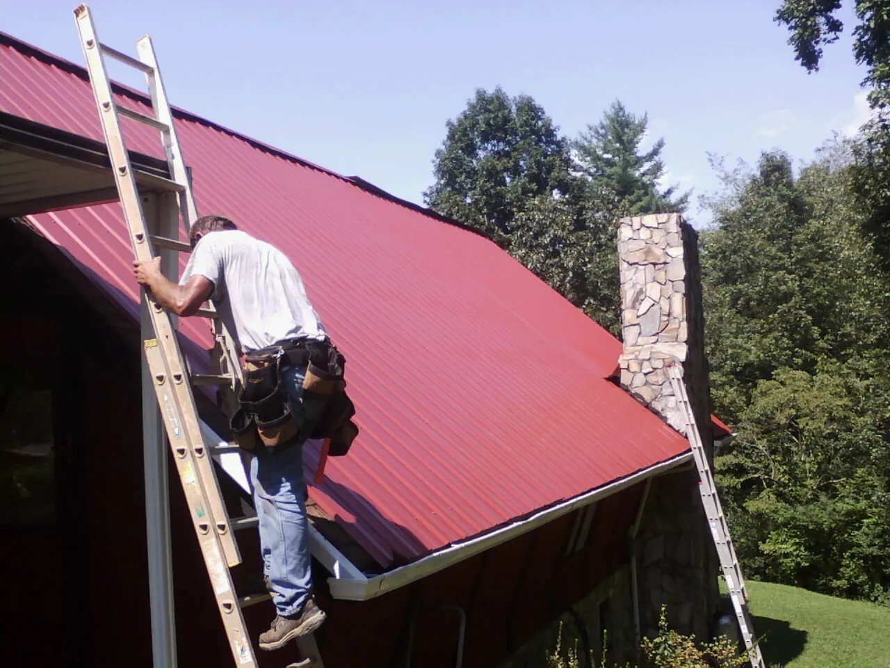 Roofer on a ladder beside a red metal roof and stone chimney — Soffit & Fascia Repair by Roofing Brothers in Boswell